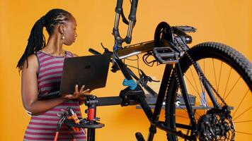 africano americano ingeniero haciendo bicicleta servicio en estudio antecedentes reparar comercio, mirando en ordenador portátil mantenimiento lista. profesional comprobación bicicleta partes ese necesitar fijación, cámara si video