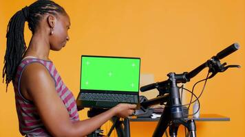 Cheerful woman following tutorial videos on chroma key laptop, learning to fix bike. BIPOC cycling hobbyist having fun repairing bicycle, following online guide on isolated screen notebook