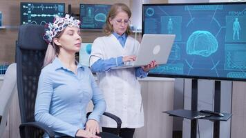 Female doctor in neuroscience checking brain data on her laptop from patient with headset full of brains sensors. video