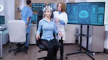 Woman wearing brainwave scanning headset sitting on a chair. Female doctor adjusting brain sensors. video