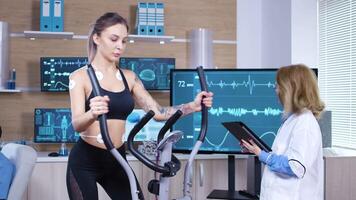 hembra atleta carreras en rueda de andar con electrodos atado en su cuerpo. hembra científico utilizando tableta a controlar electrocardiograma video