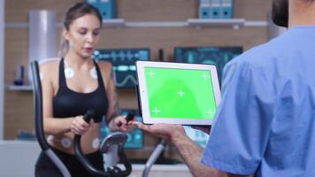 médico participación tableta con verde pantalla en frente de atleta. hembra atleta corriendo con electrodos adjunto a su cuerpo. video