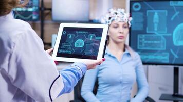 Middle age female doctor holding tablet with patient brain activity. Patient wearing brainwaves headset. video