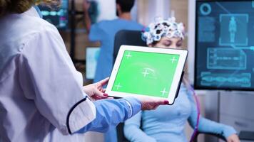 Female doctor holding tablet with green screen in a modern neurology lab science with patient sitting on a chair wearing brain waves scaning headset. video