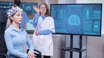 Focused female neuroscientist making hand gestures in modern facility for brain study. Female patient with brainwaves scanning headest. video