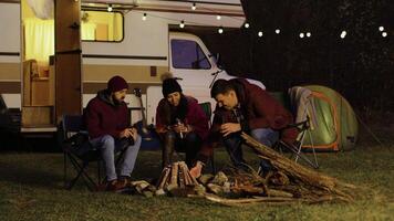 Man helping friend to light the camp fire in front of retro camper. Cold night. Light bulbs in the background. video