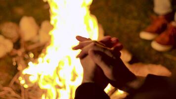 Close up of tourist sitting near the camp fire at night with his friends. Camper warming up his hands. Handheld footage video