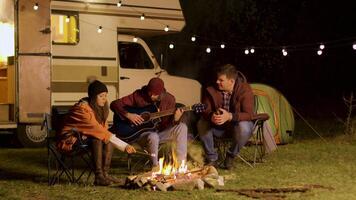 homme en chantant une chanson sur guitare pour le sien copains autour camp Feu dans une du froid nuit de l'automne dans le montagnes. rétro campeur van. lumière ampoules. video