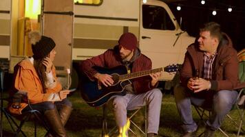 Girl holding marshmallow and one of his her friends is singing on a guitar around camp fire in cold night of autumn. Camping in mountains. video