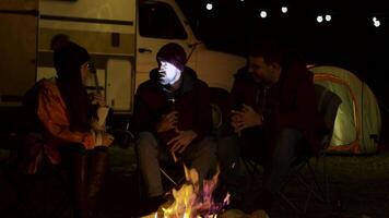 Bearded man telling a scary story to his friends around camp fire in a cold night of autumn. Retro camper van. Light bulbs video