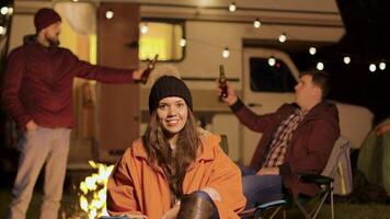 ragazza seduta su un' campeggio sedia nel un' freddo notte di autunno. amici tintinnante birra bottiglie nel il sfondo. video