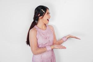 A cheerful Asian woman wearing pink dress is pointing copy space beside her, isolated by white background. photo