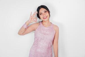 Attractive young Asian woman in pink dress is gesturing OK sign with her fingers, isolated by white background photo