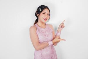 A cheerful Asian woman wearing pink dress is pointing copy space beside her, isolated by white background. photo