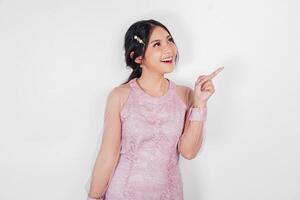 A cheerful Asian woman wearing pink dress is pointing copy space beside her, isolated by white background. photo