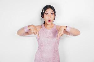Shocked Asian woman dressed in pink, pointing at the copy space below her, isolated by white background photo