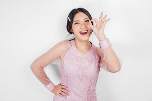 Attractive young Asian woman in pink dress is gesturing OK sign with her fingers, isolated by white background photo