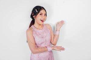 A cheerful Asian woman wearing pink dress is pointing copy space beside her, isolated by white background. photo