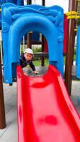 un alegre pequeño chico corredizo en un vistoso patio de recreo equipo. un pequeño chico jugando en un rojo y azul diapositiva foto