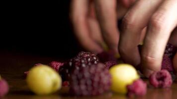 männlich Hand nimmt Beeren und hölzern Tabelle auf schwarz Hintergrund. männlich Hand nimmt Brombeere, Cranberry, Johannisbeere, Stachelbeere, Meer Sanddorn und andere Beeren fallen isoliert auf schwarz Hintergrund. video