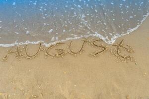 The word August is written on the sand of a beach by the sea. Washed off by a wave. The inscription disappears. End of summer concept. Back to school. photo