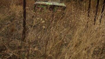 Rusty old abandoned green car lying in a tall grass field behind the fence. Shot. Wrecked rusty russian vehicle forgotten in the countryside field behind the fence. video