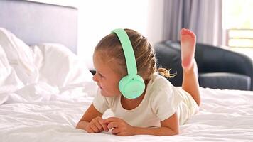 Little girl laying in bed and listening music with green kids headphones in home bedroom. High quality 4k footage video