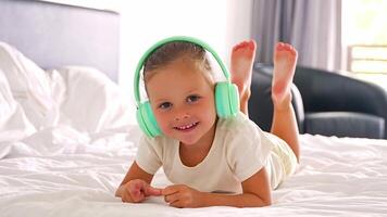 pequeño niña tendido en cama, canto y escuchando música con verde niños auriculares en hogar dormitorio. alto calidad 4k imágenes video