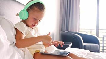 Little girl in headphones using digital tablet while listening to music or playing game in home bed. High quality 4k footage video