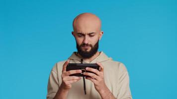 Middle eastern person playing mobile video games on app, standing over blue background and enjoying online tournament using mobile phone. Man having fun with gaming contest. Camera 2.