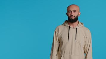 Mitte östlichen ernst Erwachsene posieren auf Studio Hintergrund, Gefühl zuversichtlich und lächelnd Über Türkis Hintergrund. Arabisch Person Schauspielkunst beiläufig im ein Beige Kapuzenpullover, gut aussehend Kerl. Kamera 1. video