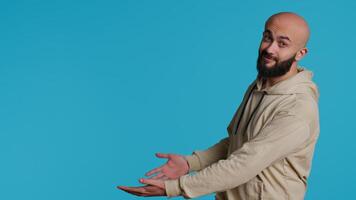 Smiling person advertising something on camera, pointing to left or right sides in studio. Middle eastern guy showing advertisement for marketing campaign, blue background pose. Camera 1. video