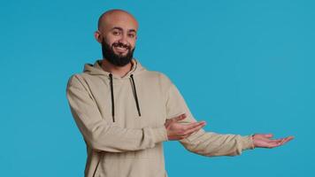 Cheerful man creating ad in front of studio camera, pointing to left or right sides over blue background. Middle eastern person showing commercial or advertisement for marketing campaign. Camera 1. video