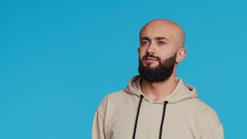 Middle eastern serious adult posing on studio backdrop, feeling confident and smiling over turquoise background. Arabic person acting casual in a beige hoodie, handsome guy. Camera 2. video