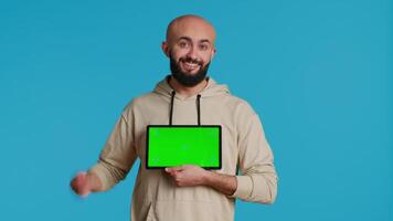 Arab guy holding device with greenscreen blank display, presenting isolated chromakey layout on tablet. Middle eastern adult using gadget to create promotional ad with copyspace. Camera 1. video