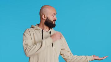 Cheerful man creating ad in front of studio camera, pointing to left or right sides over blue background. Middle eastern person showing commercial or advertisement for marketing campaign. Camera 2. video