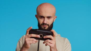 Middle eastern guy enjoying video games contest on camera, playing online mobile competition with his friends. Arab person having fun with gaming shooter tournament. Camera 2. Handheld shot.