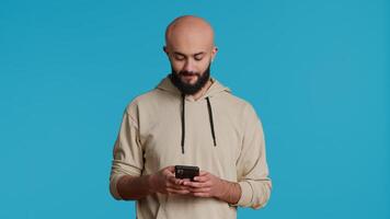 Arab person typing text messages on smartphone app, using mobile phone internet to check social media. Middle eastern guy browsing online websites in studio, blue backdrop. Camera 1. video