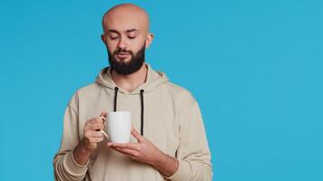 Muslim person enjoying fresh caffeine aroma in studio, drinking cup of coffee and standing over blue background. Middle eastern adult smelling beverage and taking a sip. Camera 2. Handheld shot. video