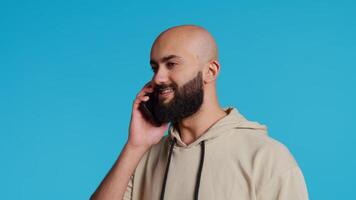 Middle eastern guy chatting with friends on phone call, answering smartphone while standing over blue background. Modern man discussing something on a remote network. Camera 2. Handheld shot. video