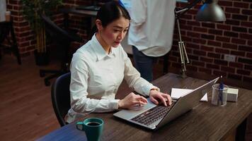 Smiling asian employee writing important emails at computer desk next to full mug of coffee. Cheerful employee typing on laptop, solving tasks, communicating with business partners video