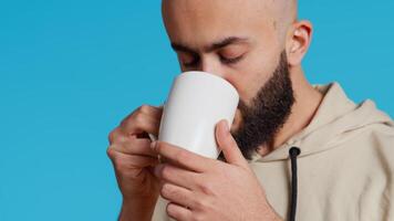 medio oriental chico Bebiendo taza de café en estudio, disfrutando Fresco aroma de cafeína refresco. árabe persona en pie terminado azul fondo, tomando un sorbo desde taza. cámara 2. Mano disparo. video