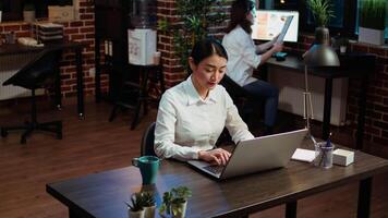 Smiling woman sitting down at office desk, typing on laptop keyboard, starting work with mug full of coffee, solving tasks late at night. Cheerful employee imputing data on computer video