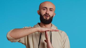 Middle eastern person doing timeout gesture on camera, asking for a break to stop working over blue background. Muslim adult presents symbol to pause or stop something. Camera 2. Handheld shot. video