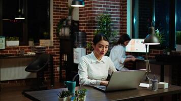 Portrait of smiling asian businesswoman doing computer tasks for team project in office. Cheerful employee working late at night, typing on laptop keyboard next to coworker, camera A video