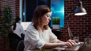 Close up of smiling businesswoman working for company project, doing tasks in brick wall office overnight. Cheerful employee inputting data on laptop late at night, zoom in shot video