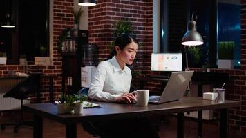 mujer de negocios transcribiendo información desde ordenador portátil a bloc, haciendo Tareas para equipo proyecto en oficina durante la noche. trabajador trabajando tarde a noche, escritura abajo datos desde computadora pantalla video