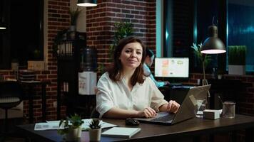 Portrait of smiling accountant researching key data for company project, doing tasks in office overnight. Happy employee sitting at desk in front of laptop, looking over accounting figures, camera A video