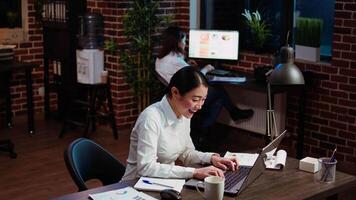 Asian woman dancing in office, feeling excited after learning of upcoming promotion, rewarded for good results. Cheerful employee singing on computer desk chair, celebrating, camera A video