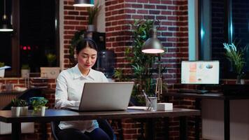 sorridente asiatico lavoratore ricercando chiave dati per azienda progetto, fare compiti nel ufficio durante la notte. allegro personale membro guardare al di sopra di contabilità figure su il computer portatile schermo in ritardo a notte, digitando su tastiera video